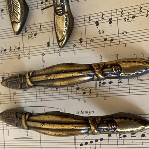 May include: A collection of metallic brass-toned drawer pulls featuring a textured, elongated design with pointed ends and a central wrapped detail. The hardware has a weathered finish with dark patina accents along the ridges and grooves.