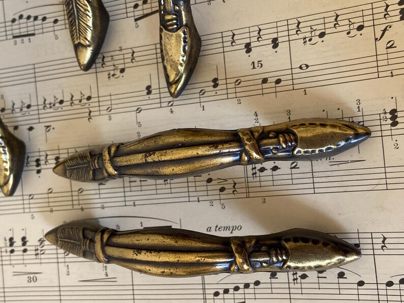 May include: A collection of metallic brass-toned drawer pulls featuring a textured, elongated design with pointed ends and a central wrapped detail. The hardware has a weathered finish with dark patina accents along the ridges and grooves.