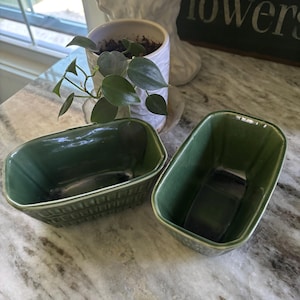 May include: Two rectangular ceramic loaf pans with a glossy dark green glaze. Each pan features a molded grid pattern on the exterior sides and a smooth interior finish. The vessels have rounded corners and a slightly flared rim design.