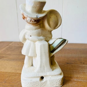 May include: A white ceramic figurine of a person wearing a top hat and holding a bag. The figurine is standing on a base with a textured surface.