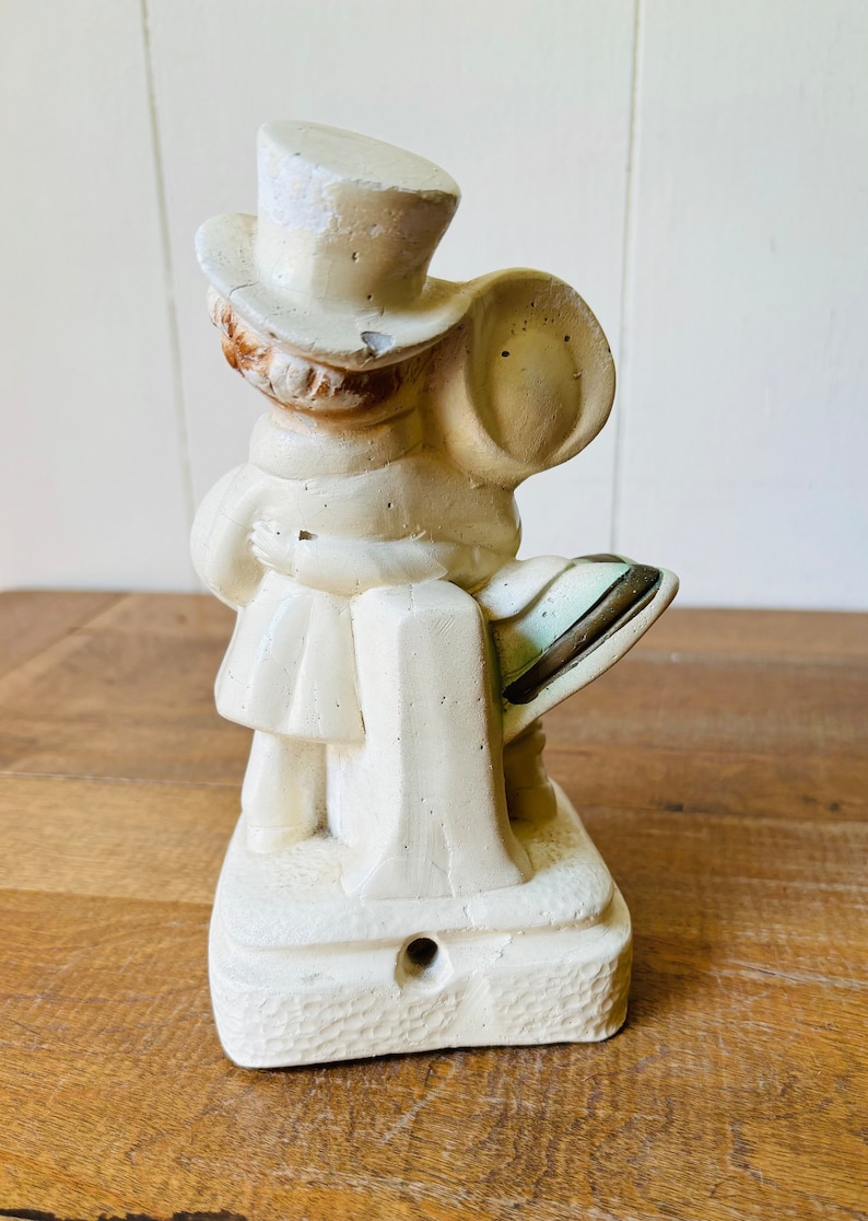May include: A white ceramic figurine of a person wearing a top hat and holding a bag. The figurine is standing on a base with a textured surface.