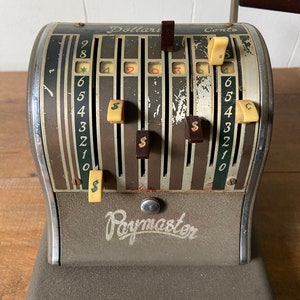 May include: A vintage Paymaster cash register with a brown and silver metal body. The register has a series of dials with numbers 1-9 and a dollar sign. The register has a wooden handle.