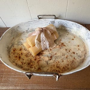 May include: An oval, distressed white metal tray with handles, holding several light-colored bars tied with a pink ribbon and lace. The tray has a rustic, aged appearance, with brown speckling on the surface.