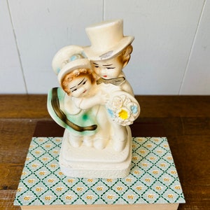 May include: A vintage ceramic figurine of a bride and groom holding a bouquet of flowers. The figurine is painted in white, blue, and yellow.