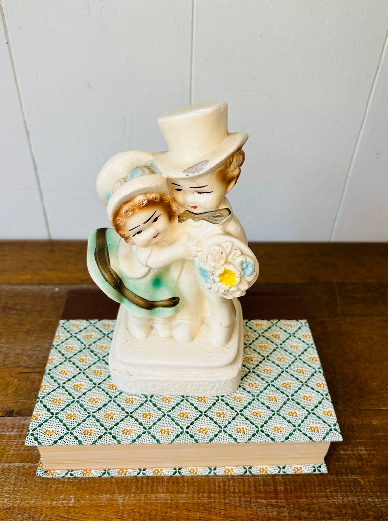 May include: A vintage ceramic figurine of a bride and groom holding a bouquet of flowers. The figurine is painted in white, blue, and yellow.