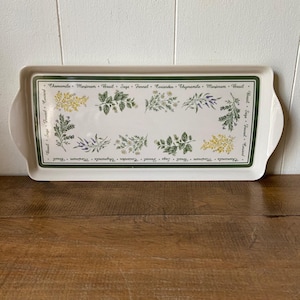 May include: A rectangular white ceramic serving tray with integrated handles. The center features a botanical illustration of various herbs and flowers, surrounded by a dark green border printed with the names Chamomile, Marjoram, Basil, Sage, Fennel, and Coriander.