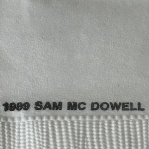 May include: Close-up of a white textile with the text "1989 SAM MC DOWELL" printed in black. The textile has a textured surface and a woven pattern at the bottom.