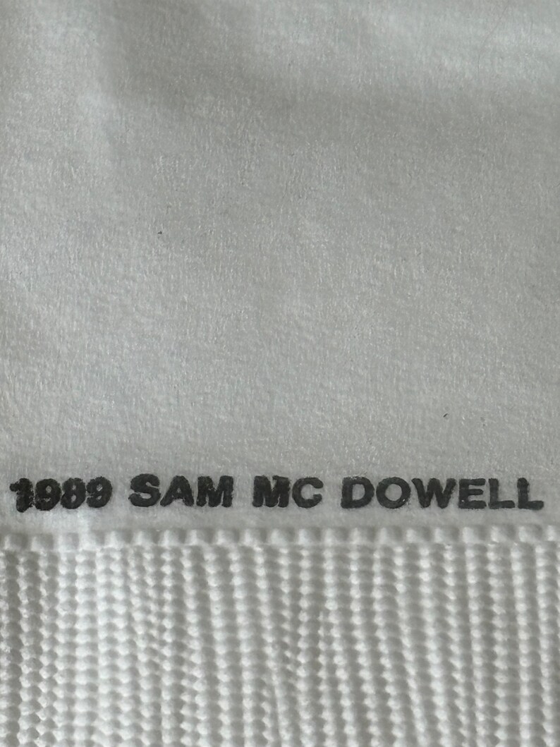 May include: Close-up of a white textile with the text "1989 SAM MC DOWELL" printed in black. The textile has a textured surface and a woven pattern at the bottom.