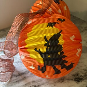 May include: Orange and yellow paper Halloween lanterns with a witch silhouette and bat cutouts. The lanterns have a pleated design and are accented with a copper-colored mesh ribbon. Festive decor for Halloween.