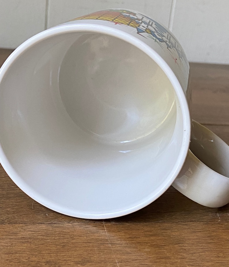 Puede incluir: Taza de cer&aacute;mica blanca con un dise&ntilde;o colorido de un edificio con un techo amarillo y ventanas azules. La taza est&aacute; vac&iacute;a y el interior es blanco.