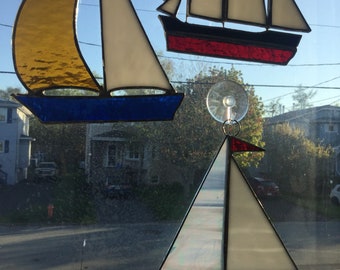 May include: Three stained glass sailboats hanging in a window. The sailboats are blue, red and white with black accents. The sailboats are suspended by suction cups.