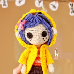 May include: A handmade crochet doll with blue hair, wearing a yellow jacket and a brown skirt. The doll has button eyes and a pink flower in its hair. A black cat illustration is next to the doll.