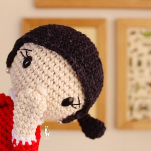 May include: A crocheted doll with black hair, a white face, and black eyes. The doll is wearing a red and white dress.