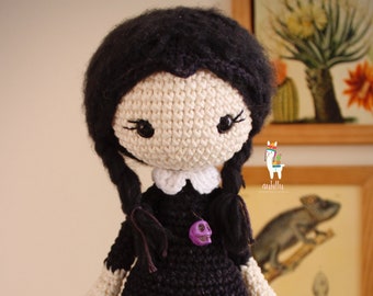 May include: Crocheted doll with black hair, a white dress with a black collar, and a purple skull charm. The doll is holding a black crocheted cat.