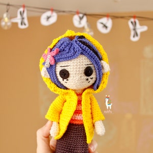 May include: A crocheted doll wearing a yellow raincoat with a purple flower on the hood. The doll has blue hair, brown eyes, and a brown skirt. The doll is holding a black cat with blue eyes.