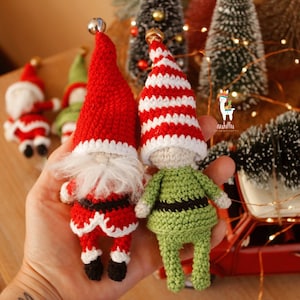 Op de afbeelding: Twee gehaakte gnome-versieringen, een in rood en wit met een witte baard en de andere in groen met een zwarte riem. De gnomes houden elkaar vast en staan voor een rode speelgoedauto.
