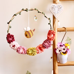 May include: A crocheted flower garland with a bee hanging from a wooden hoop. The garland is made of pink, red, and yellow flowers. The bee is yellow and black with white wings.