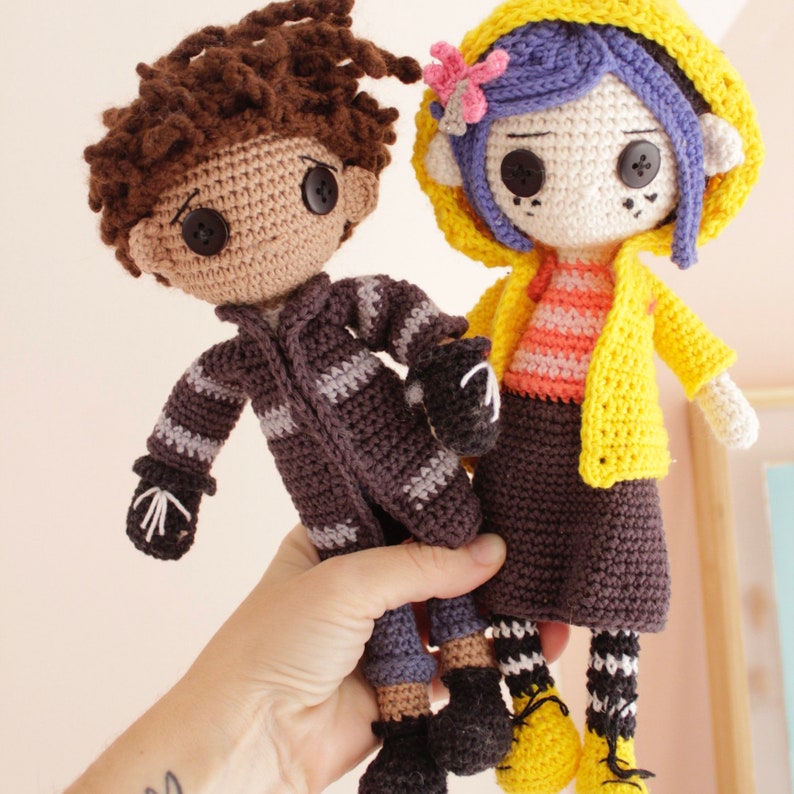 May include: Two crocheted dolls, one with brown hair and a striped jacket, the other with blue hair and a yellow raincoat.