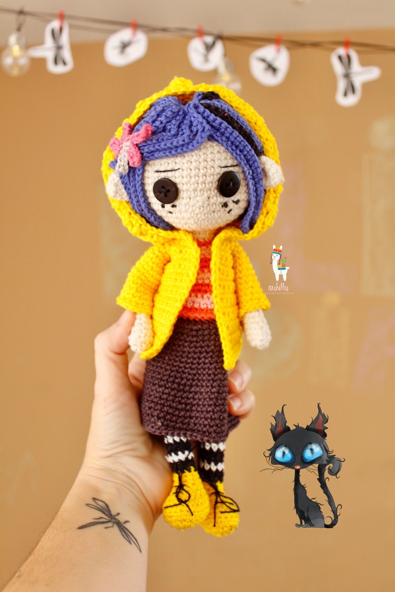 May include: A crocheted doll wearing a yellow raincoat with a purple flower on the hood. The doll has blue hair, brown eyes, and a brown skirt. The doll is holding a black cat with blue eyes.