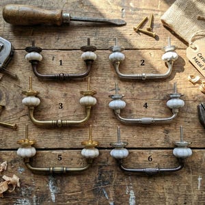 Könnte beinhalten: Sechs Vintage-Schrankgriffe aus Keramik und Metall mit weißen Keramikdetails, präsentiert auf einer Holzoberfläche. Werkzeuge und Hardware sind ebenfalls vorhanden, zusammen mit einem Etikett "Vintage Ceramic & Metal Pulls".