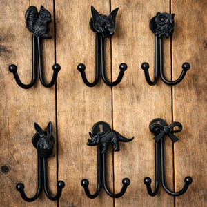 Op de afbeelding: Zes zwarte metalen wandhaken met dieren- en strikdesigns. De haken hebben een eekhoorn, vos, uil, konijn, dinosaurus en strik. Elke haak heeft twee onderste haken en een decoratieve bovenkant. De haken zijn op een houten oppervlak gemonteerd.