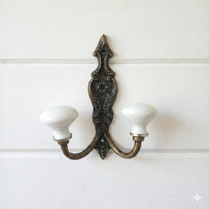 May include: Ornate, antique-style wall hook with two white ceramic knobs. The hook has a dark bronze finish with decorative floral detailing. The knobs are attached to curved arms, creating a functional and decorative piece for any room.