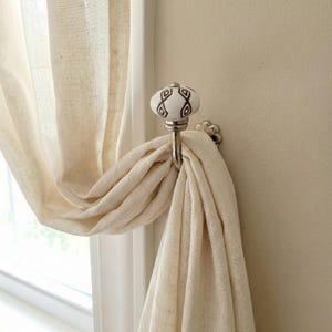 Pode incluir: Um gancho decorativo para cortina com um botão de cerâmica branco e castanho padronizado. O gancho é feito de metal prateado e segura uma cortina bege. O gancho é montado numa parede bege.