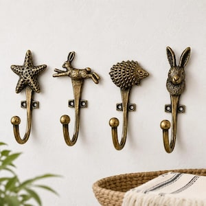 Op de afbeelding: Vier antieke messing wandhaken met dierontwerpen: een zeester, een rennende konijn, een egel en een konijnenkop. Elke haak heeft een gebogen onderkant en een ronde knop. De haken zijn aan een witte muur bevestigd.