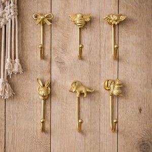 May include: Six decorative gold-coloured metal wall hooks. The hooks feature various designs, including a bow, a bee, a flower, a rabbit, an elephant, and a squirrel. The hooks are mounted on a light brown wooden surface.