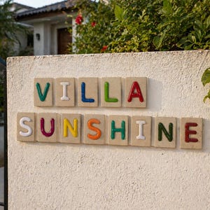 Puede incluir: Un muro de estuco blanco muestra las palabras "VILLA SUNSHINE" en letras coloridas en relieve. Las letras están colocadas sobre baldosas beige rectangulares individuales. El muro está enmarcado por follaje verde.