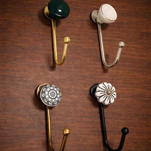 May include: Four decorative wall hooks with different designs. One has a dark green knob and gold finish, another a white knob with a silver finish, a third with a black and white floral design and gold finish, and the last with a white flower design and black finish.