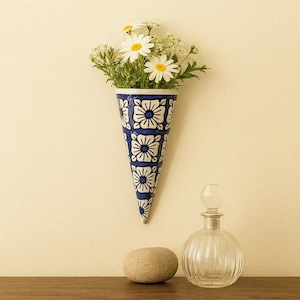 May include: A blue and white patterned cone-shaped wall vase filled with white daisies and greenery. A clear, round glass bottle with a stopper and a smooth, oval stone are on a wooden surface below.