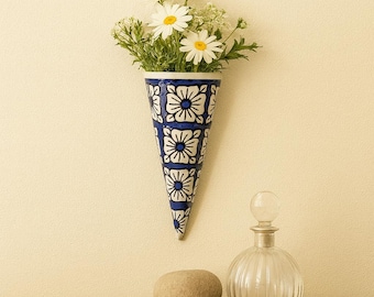May include: A blue and white patterned cone-shaped wall vase filled with white daisies and greenery. A clear, round glass bottle with a stopper and a smooth, oval stone are on a wooden surface below.