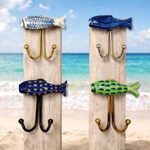 Puede incluir: Cuatro percheros de pared de madera con diseños decorativos de peces. Los peces están pintados en azul, blanco y verde con ganchos de metal dorado y negro. El fondo muestra una escena de playa.