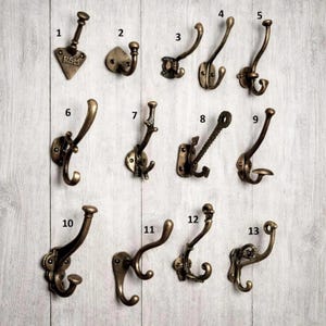 Könnte beinhalten: Eine Sammlung von dreizehn antiken, bronzefarbenen Garderobenhaken, jeder mit einem einzigartigen Design. Einige Haken haben dekorative Elemente, während andere schlicht sind. Die Haken sind an einer hellen Wand mit Holzmaserung befestigt.