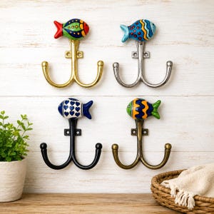 Puede incluir: Cuatro ganchos de pared con remates de peces de cerámica en varios colores y patrones. Los ganchos tienen acabados metálicos en oro, plata, negro y bronce antiguo. Cada gancho tiene dos brazos curvos y una placa de montaje con dos orificios.