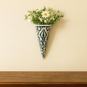 May include: A blue and white patterned, cone-shaped wall vase filled with daisies and greenery. The vase is hanging on a cream-colored wall above a wooden surface. The vase has a detailed floral design.