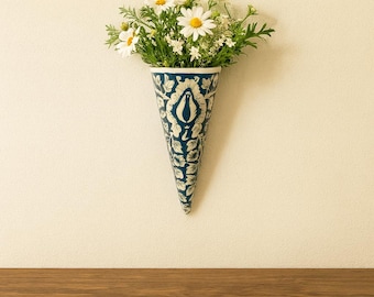 May include: A blue and white patterned, cone-shaped wall vase filled with daisies and greenery. The vase is hanging on a cream-colored wall above a wooden surface. The vase has a detailed floral design.