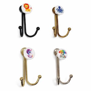 Könnte beinhalten: Vier dekorative Wandhaken mit Tiermotiven. Die Haken haben einen Metallrahmen und einen Keramikknopf mit einem Löwen, einem Oktopus, einem Einhorn und einem Einhorn mit einem Regenschirm. Die Haken sind in Schwarz, Gold und Silber gehalten.