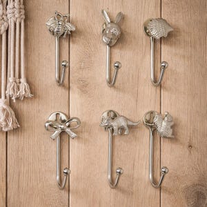 Op de afbeelding: Een verzameling zilverkleurige metalen wandhaken met decoratieve dieren- en objectontwerpen. De haken omvatten een bij, konijn, egel, strik, dinosaurus en eekhoorn. De haken zijn gemonteerd op een lichtbruin houten oppervlak.