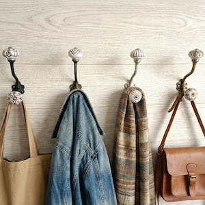 May include: Four decorative wall hooks with ceramic knobs and ornate metalwork. A tan tote bag, a denim jacket, a patterned scarf, and a brown leather satchel hang from the hooks, showcasing a rustic, farmhouse style.