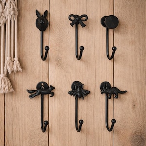 May include: Six black metal wall hooks with decorative animal and bow designs. The hooks include a rabbit, bow, circle, fox, bee, and dinosaur. The hooks are mounted on a light brown wooden surface.
