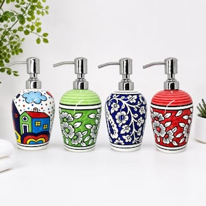 May include: Four ceramic soap dispensers with silver pump tops. The dispensers are painted in various designs: a house scene, green floral, blue floral, and red floral. Each dispenser is approximately 15 cm tall.
