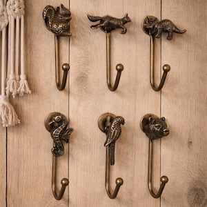 May include: A collection of six bronze-toned metal wall hooks, each with a different animal design. The hooks include a squirrel, fox, rhinoceros, seahorse, parrot, and bear. They are mounted on a light brown wooden surface.