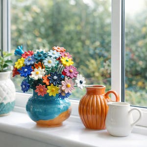 Peut inclure: Un vase en céramique bleu avec un bouquet de fleurs en forme de marguerites multicolores. À côté, un pichet orange et un mug blanc, le tout sur un rebord de fenêtre. En arrière-plan, une vue floue d'arbres et de feuillage verts.
