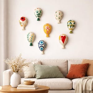 May include: Eight ceramic hot air balloon wall decorations, each with a unique hand-painted design. The designs include floral patterns, a heart, and an evil eye. The balloons are mounted on a white wall above a sofa, creating a whimsical display.