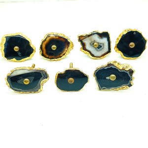 May include: A collection of seven agate cabinet knobs with gold-colored edges and brass hardware. The knobs feature a variety of colors, including black, brown, white, and gray, and are set against a white background.