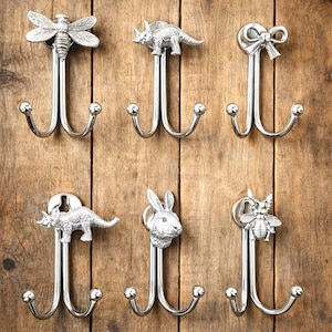 May include: A collection of six silver-colored metal wall hooks. Each hook features a decorative top: a dragonfly, a dinosaur, a bow, a second dinosaur, a rabbit, and a bee. The hooks have a double-pronged design.