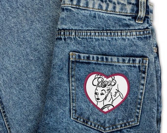 Op de afbeelding: Blauwe denim jeans met een hartvormige patch op de achterzak. De patch heeft een zwart-witte illustratie van een vrouw met een hart en het woord "okay" erin.