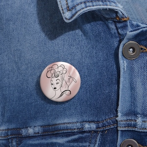 May include: A pink button with a black and white line drawing of a woman with a large hairdo. The text "Lucy" is written in cursive on the button.